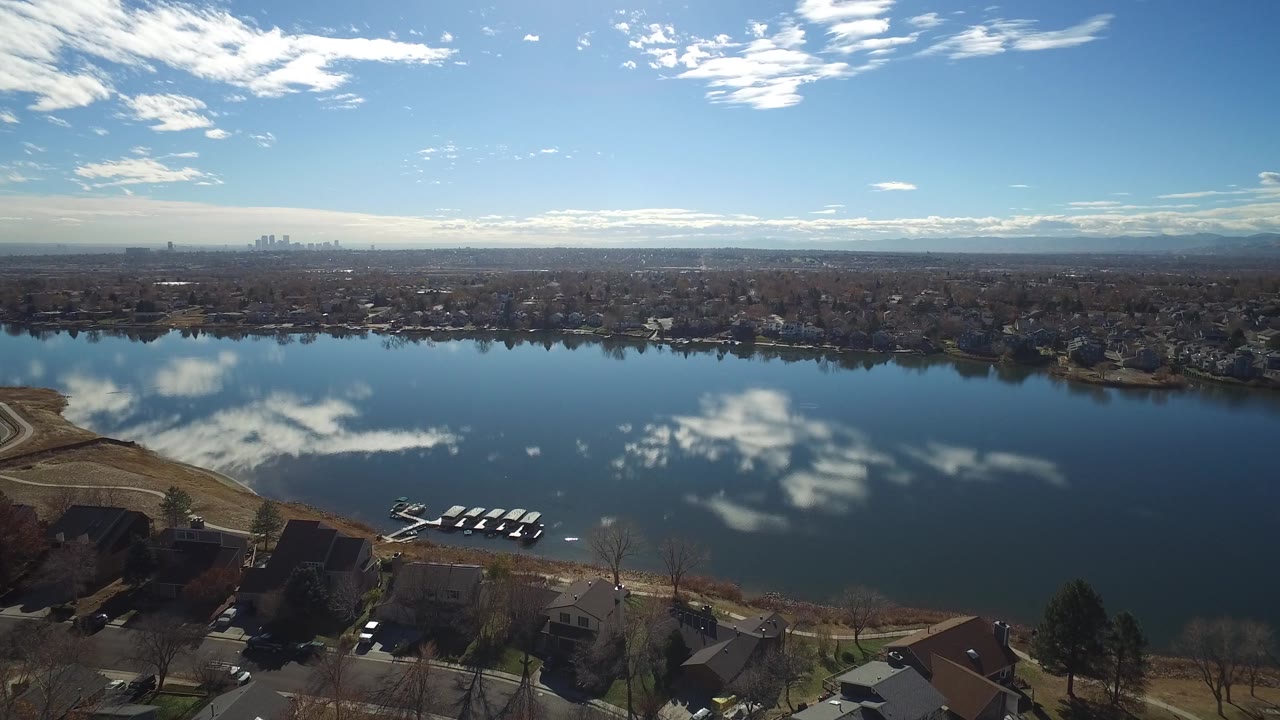 Aerial view in Westminster, Colorado with the dock lower in frame, calm lake reflections across the center, houses along the far shore, and no visible people.