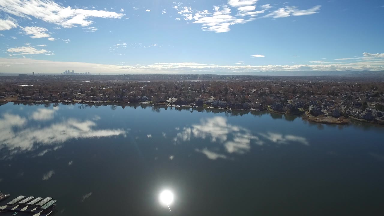 Wide aerial view over a lake toward a residential shoreline and distant skyline.