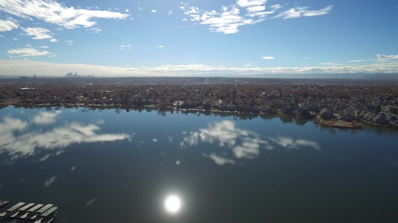 Second aerial moment over the Westminster, Colorado lake, with the sun glare lower in the water, cloud reflections across the surface, shoreline homes, bare trees, and no visible people.