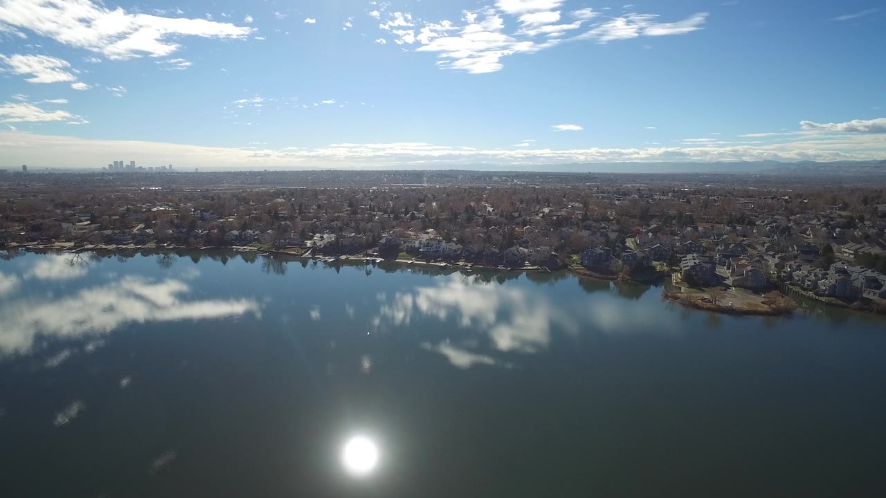 Wide aerial view over a lake with sun glare, cloud reflections, shoreline homes, distant skyline, and mountains.