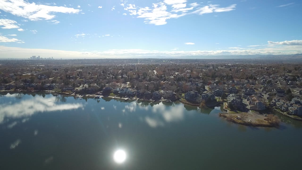 Aerial frame in Westminster, Colorado looking across a lake with a bright sun reflection, shoreline homes, distant skyline, mountain horizon, and no visible people.