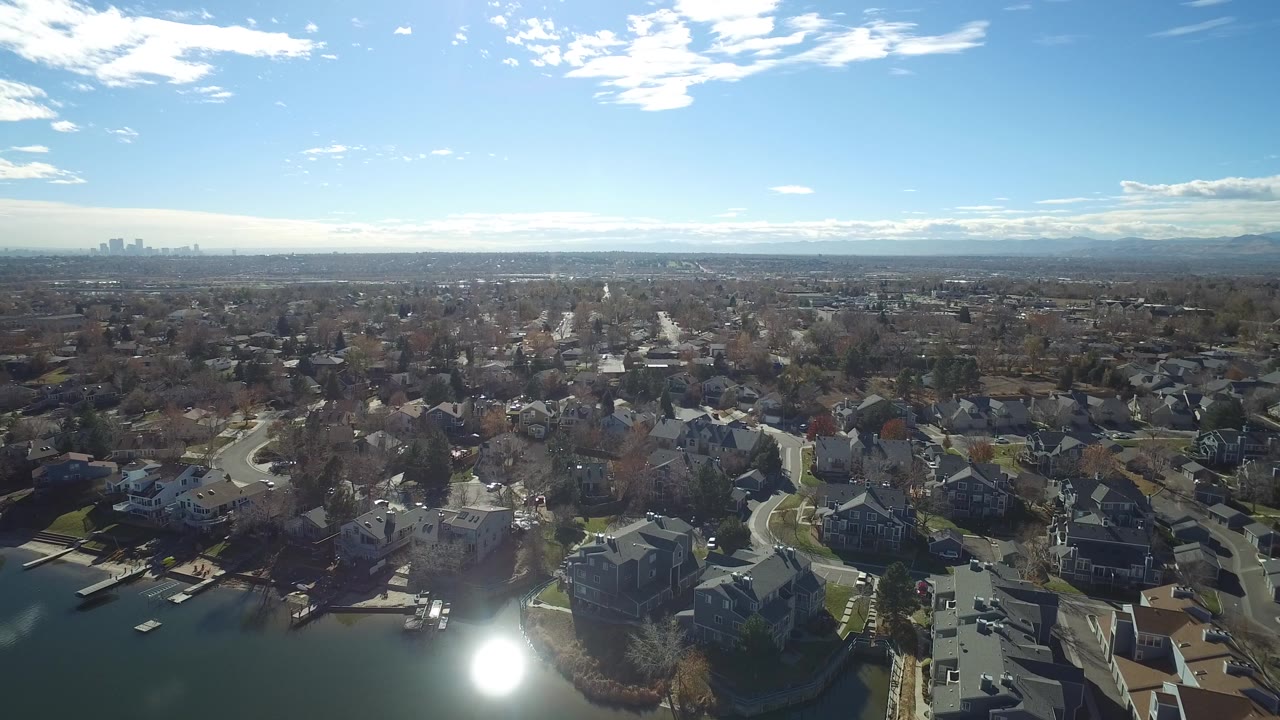 High aerial view over Westminster, Colorado homes, curving streets, a lake edge, distant skyline, and mountain horizon, with bright sun glare and no visible people.
