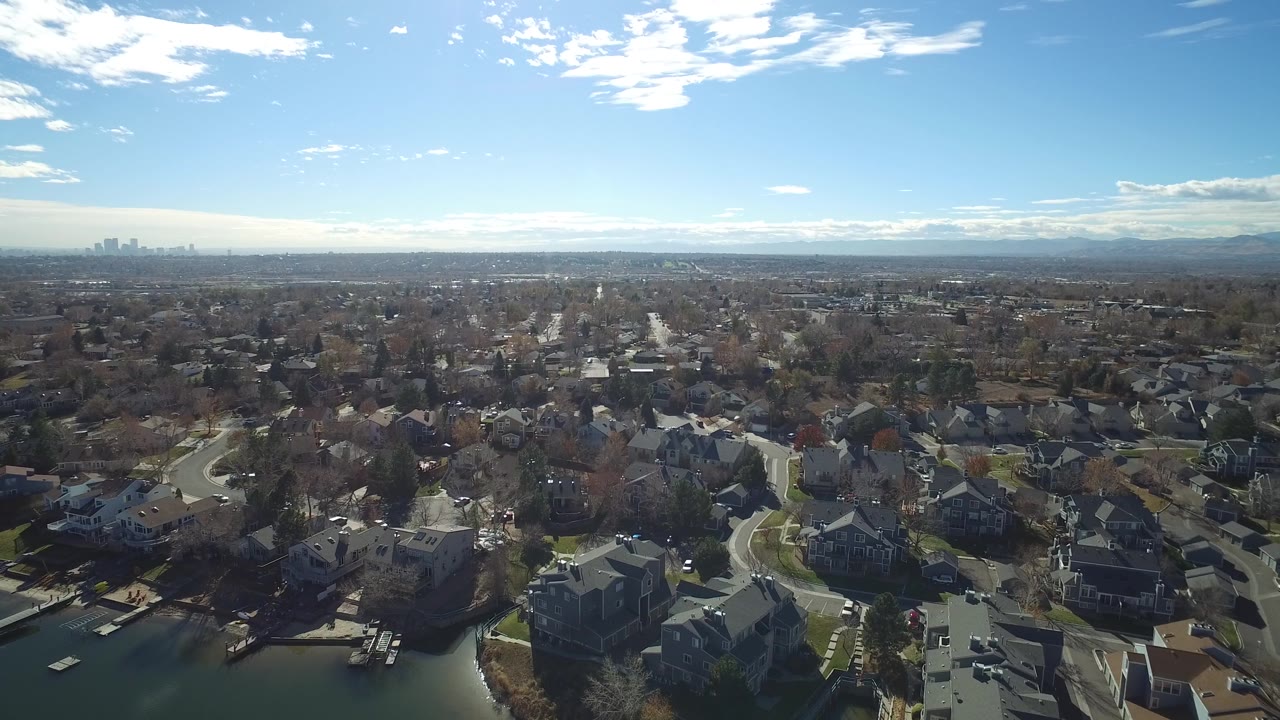 Wide aerial panorama of suburban blocks with homes, bare trees, skyline, and mountains.