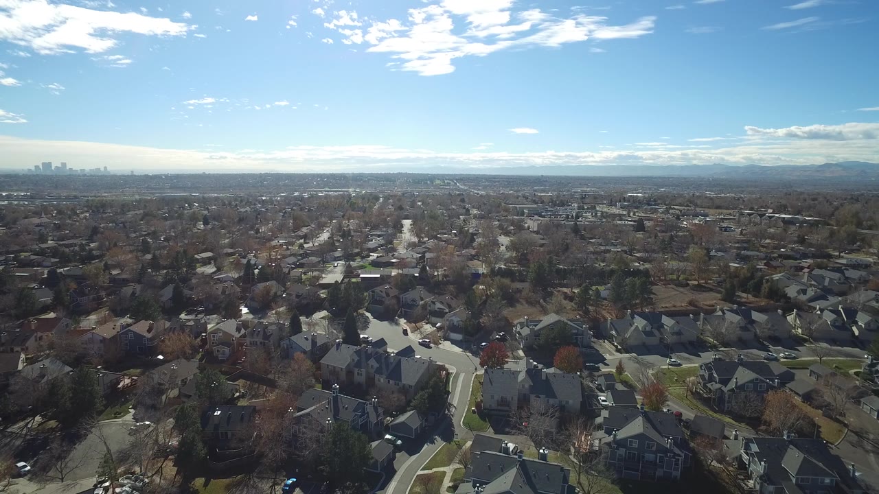 Aerial frame over Westminster, Colorado rooftops and curved streets, with a hazy city skyline left of center and no visible people.