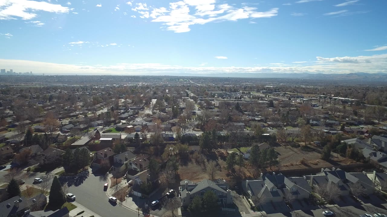 Opening aerial view over Westminster, Colorado residential blocks, leafless trees, distant skyline, mountain horizon, and bright blue sky, with no visible people.