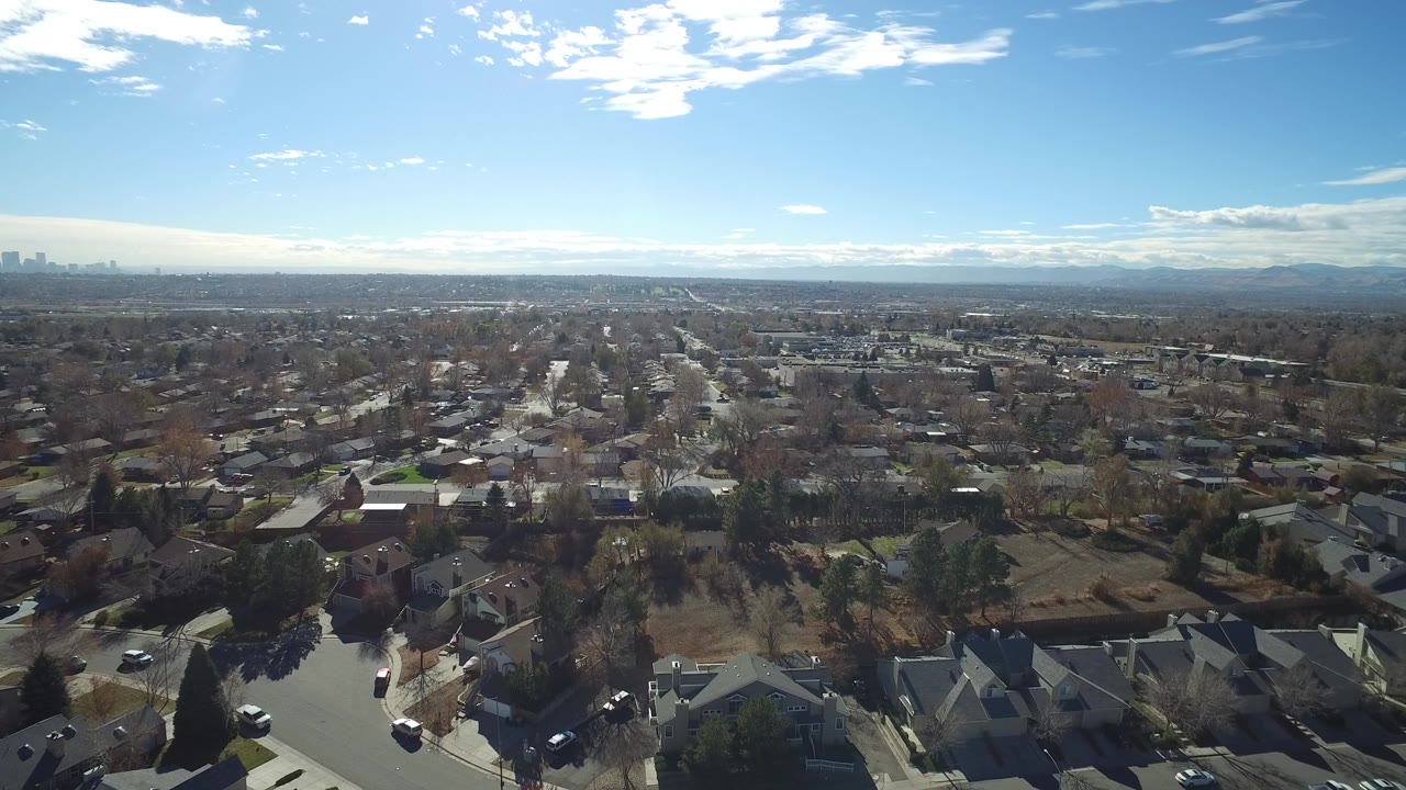 Early clip frame looking across Westminster, Colorado rooftops and tree-lined streets toward a far city skyline and hazy mountains under scattered clouds, with no visible people.