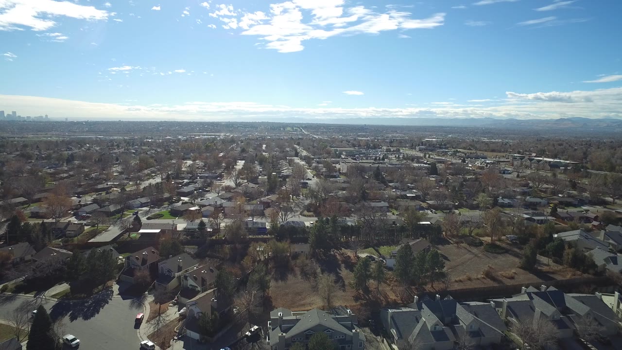 Wide suburban aerial with houses, trees, roads, and a low mountain horizon.