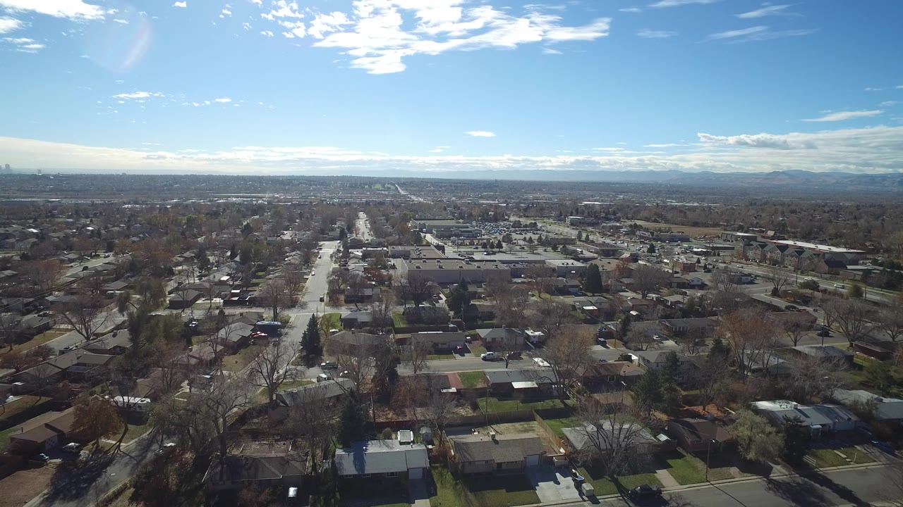 Opening aerial frame over Westminster, Colorado neighborhoods with single-story homes, bare trees, a straight street through the center, distant mountains, and no visible people.
