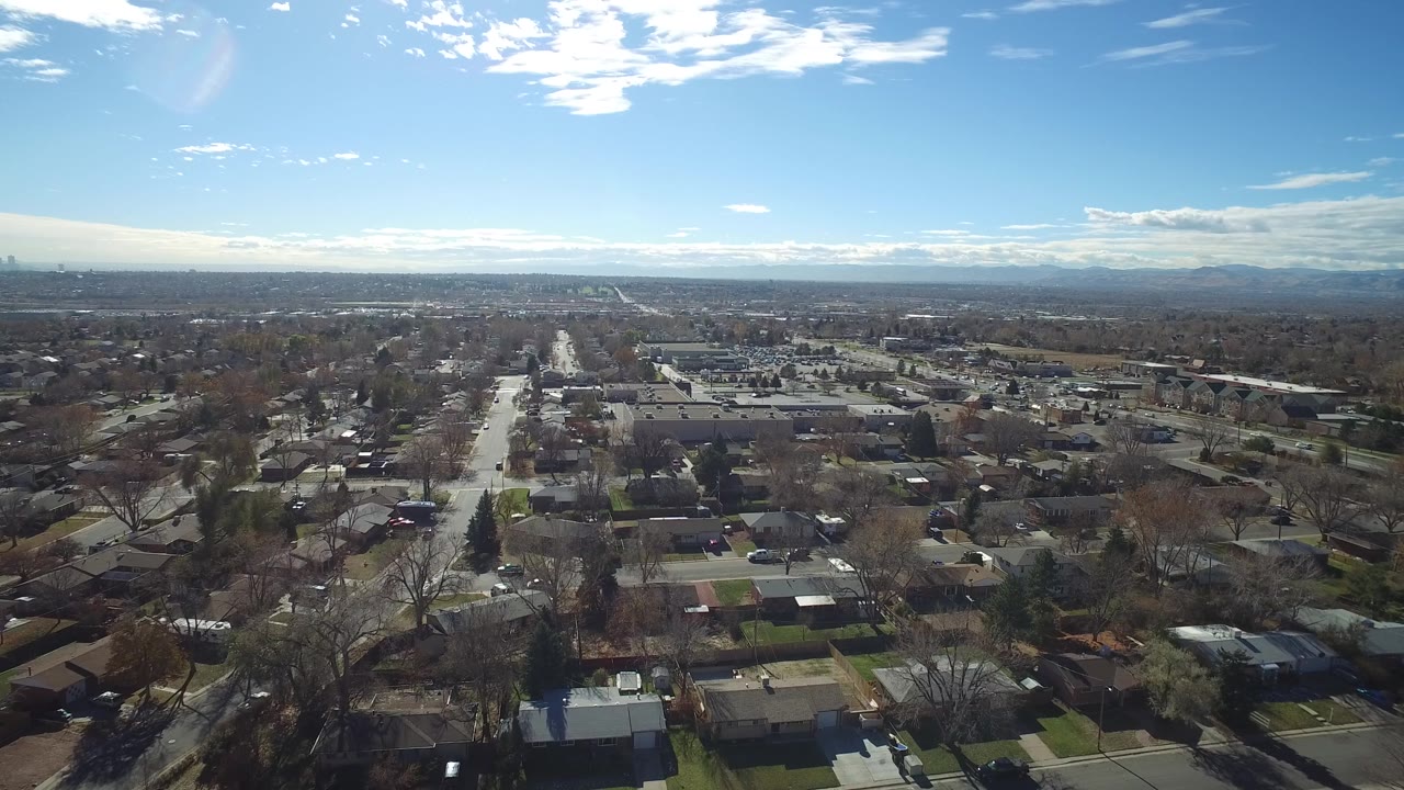 Westminster, Colorado aerial view with rooftops and yards in the foreground, a bright central street, low buildings beyond, mountains on the horizon, and no visible people.