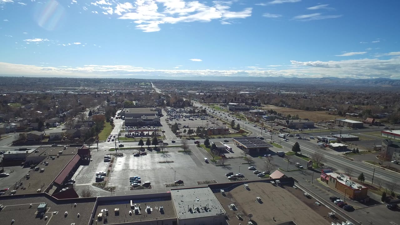 Aerial frame over Westminster, Colorado showing a large reflective parking lot, nearby roads, low buildings, distant suburbs, mountains, and no visible people.