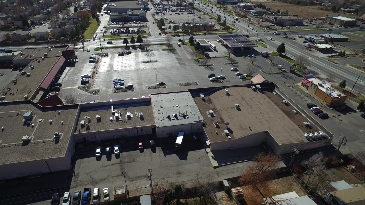 A high aerial frame over Westminster, Colorado shows flat-roof buildings, parking lots, a diagonal multi-lane road, distant businesses, and no visible people.