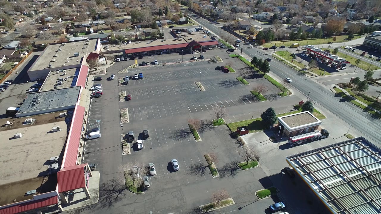 Westminster, Colorado aerial frame with the retail parking lot centered, a road along the right side, residential homes beyond the stores, and no visible people.