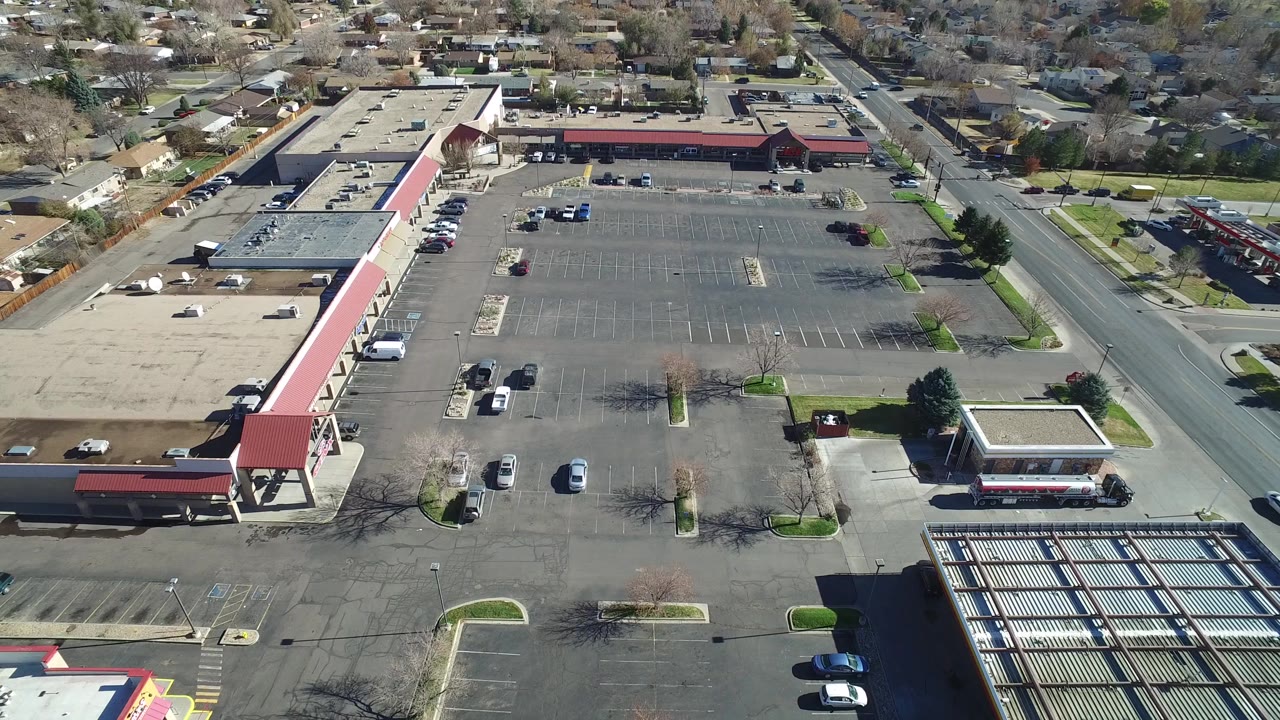 Aerial view in Westminster, Colorado with the mostly empty parking lot centered, commercial roofs along the left and top, a straight road at right, and no visible people.