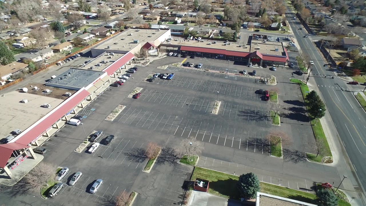 Opening aerial frame over a mostly empty parking lot in Westminster, Colorado, with red-roofed storefronts, parked cars, a multi-lane road, nearby houses, and no visible people.