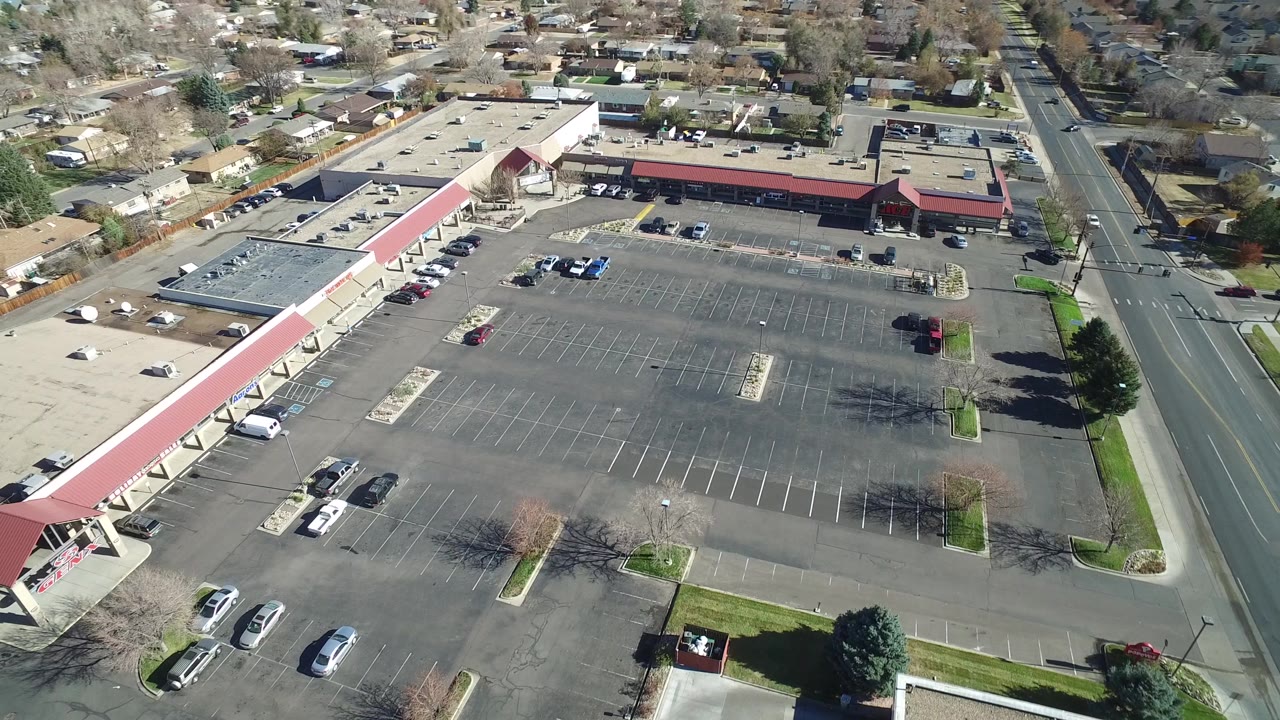 Second aerial frame in Westminster, Colorado, showing the broad asphalt lot, storefront row, scattered parked vehicles, residential blocks beyond, and no visible people.