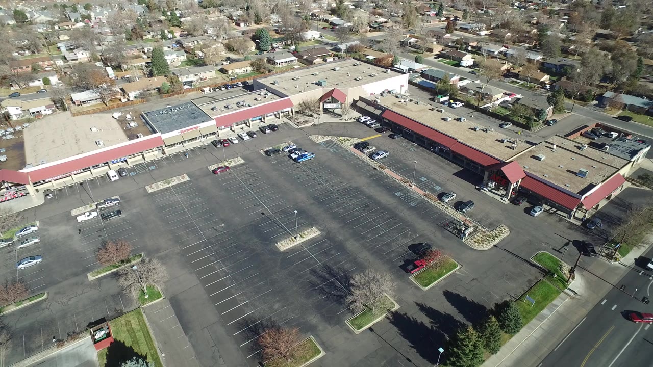 A high aerial view in Westminster, Colorado shows a broad asphalt parking lot beside red-roofed retail buildings, with roads and houses beyond and no visible people.