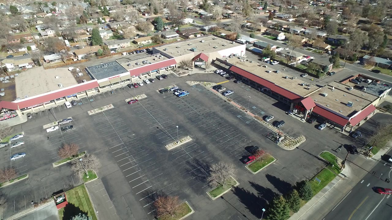 The Westminster, Colorado aerial angle holds on the strip mall and expansive parking area, with a few vehicles, bare trees, and residential blocks in the background and no visible people.