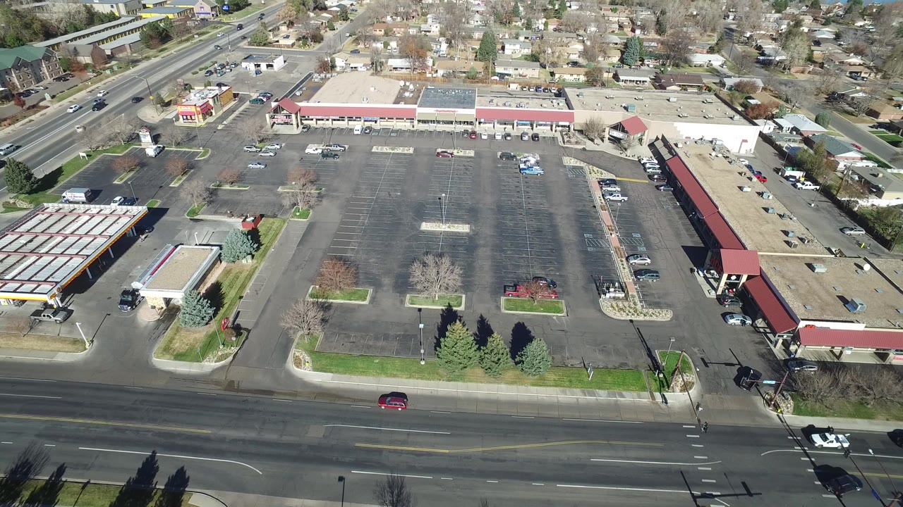 Aerial frame over Westminster, Colorado showing a wide road in front of a mostly empty commercial parking lot, a gas station canopy, low buildings, nearby homes, and no clearly visible identifiable people.