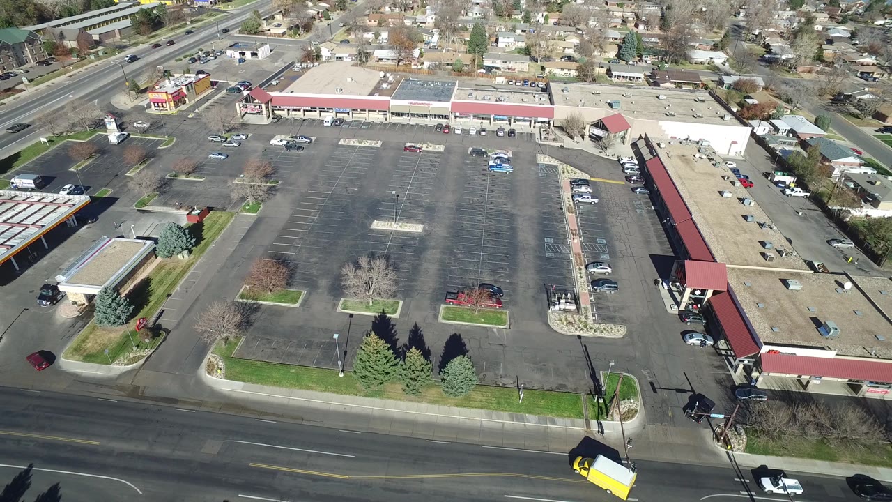 The next aerial frame keeps the Westminster strip mall centered, with empty parking rows, a few parked cars, a foreground roadway, and no visible people.