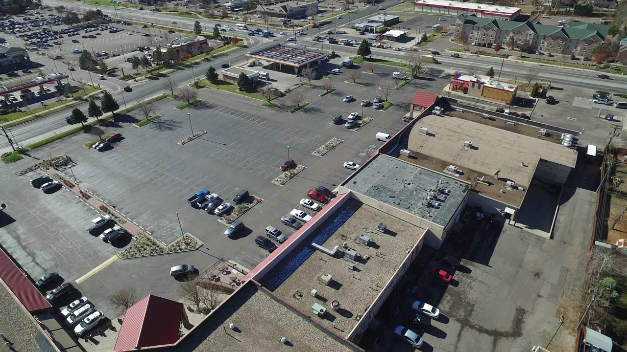 Aerial frame in Westminster, Colorado showing an open parking lot, low commercial rooftops, scattered parked cars, and cross traffic near the top of the frame with no visible people.