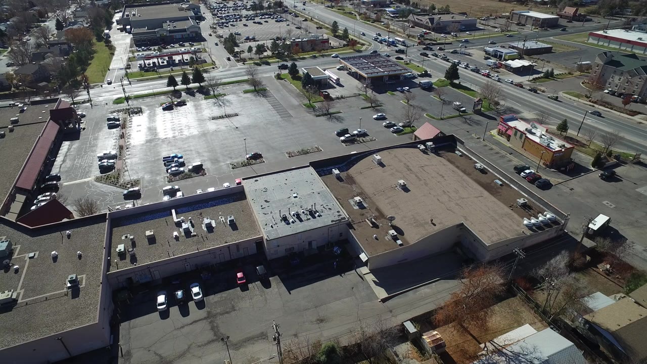 Aerial view in Westminster, Colorado over flat-roof commercial buildings, a broad parking lot, streets, parked cars, nearby houses, and no visible people.