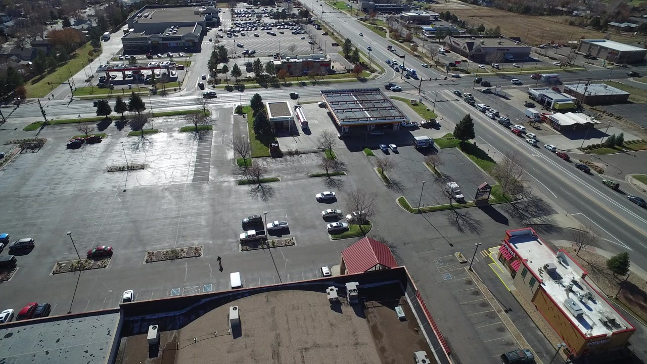Aerial frame over Westminster, Colorado showing a large reflective parking lot, multi-lane roads, low commercial buildings, and no clearly visible people.