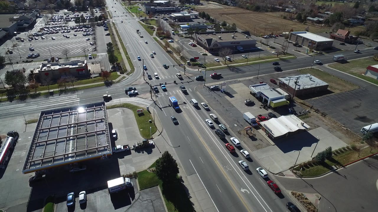 Aerial view over Westminster, Colorado showing a wide multi-lane road, gas station canopies, low commercial buildings, parking lots, grassy strips, and no clearly visible people.