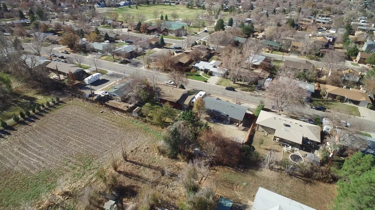 Aerial view over Westminster, Colorado with beige and gray rooftops, fenced yards, a field edge in the foreground, intersecting streets, and no visible people.
