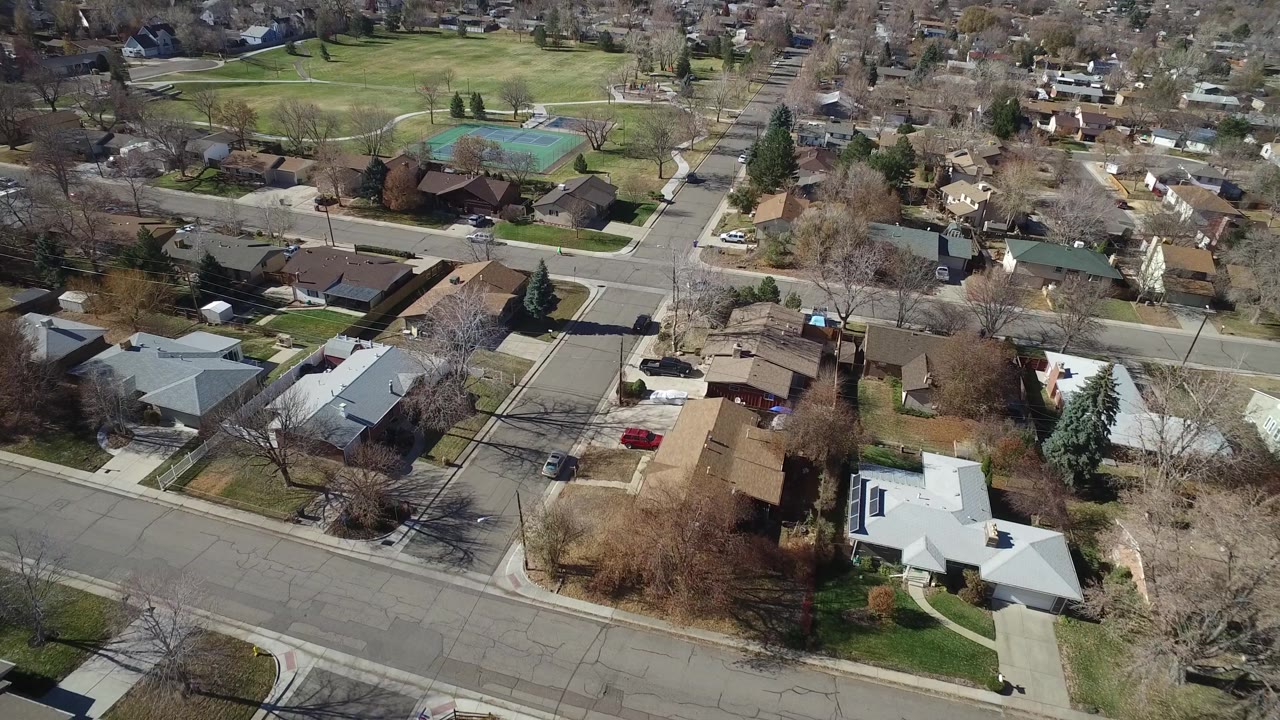 Elevated Westminster, Colorado view with rooftops and driveways in the lower frame, a cross street through the middle, and park lawns with tennis courts beyond, with no visible people.