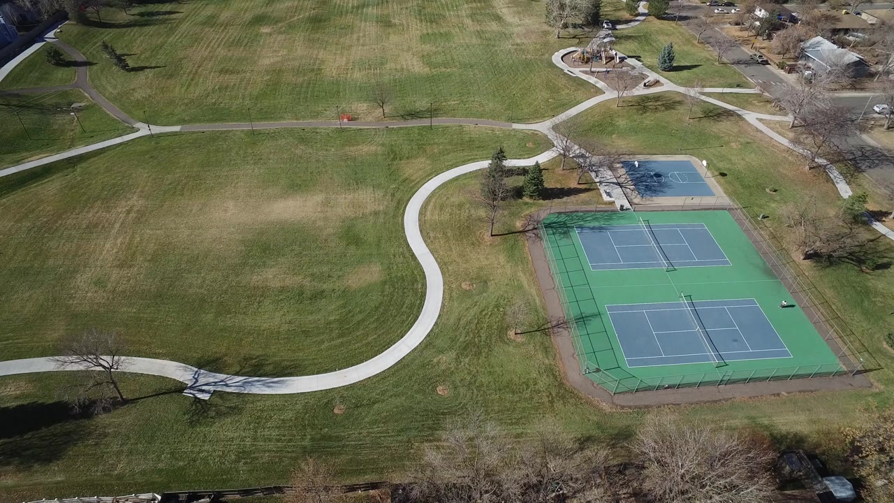 Aerial frame from Westminster, Colorado showing tennis courts slightly right of center, a pale curved path through the grass, a small blue court area beyond, houses near the top edge, and no visible people.