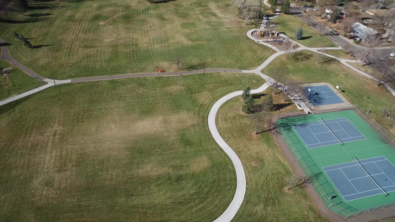 High aerial view over a Westminster, Colorado park with open grass, curved walkways, fenced tennis courts at right, nearby homes at top, and no visible people.