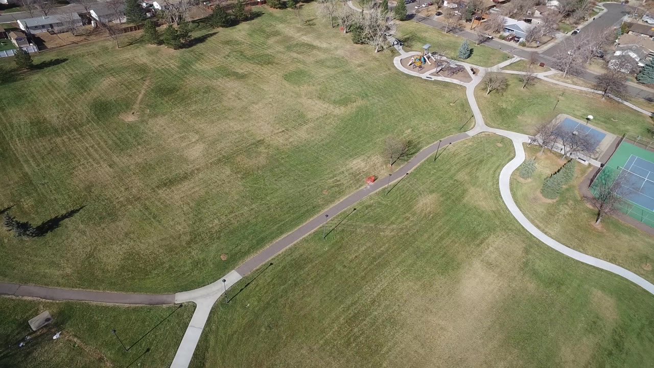 Aerial frame in Westminster, Colorado showing mowed park grass, a pale curved sidewalk, a dark path across the lower third, tennis courts at the right edge, and no visible people.