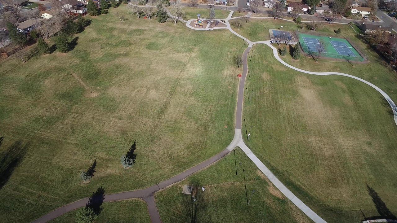 Aerial view of grass field, paths, trees, tennis courts, and nearby houses.