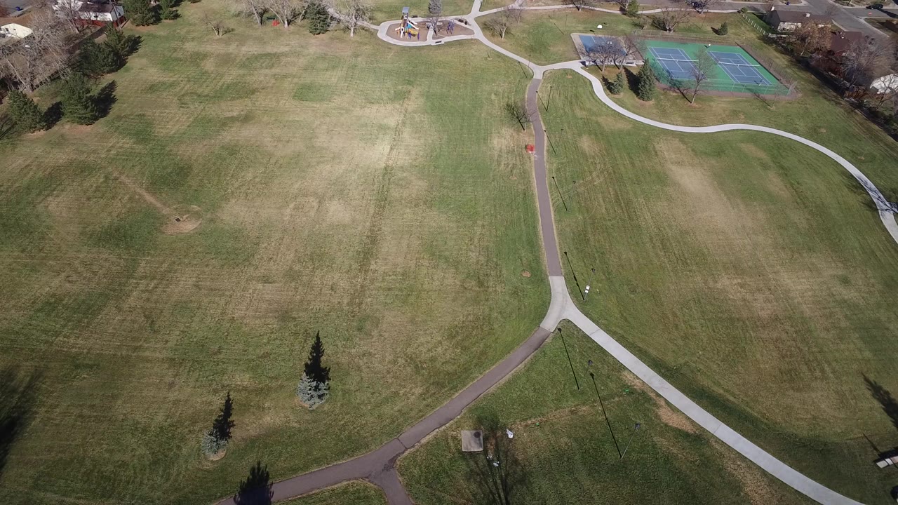 Aerial frame over Westminster, Colorado showing a broad grass field, curving paved paths, tennis courts near the upper edge, nearby houses, and no visible people.