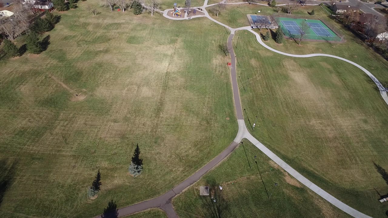 Aerial view in Westminster, Colorado with the lawn centered, intersecting paths below, tennis courts and a small playground near the top, and no visible people.