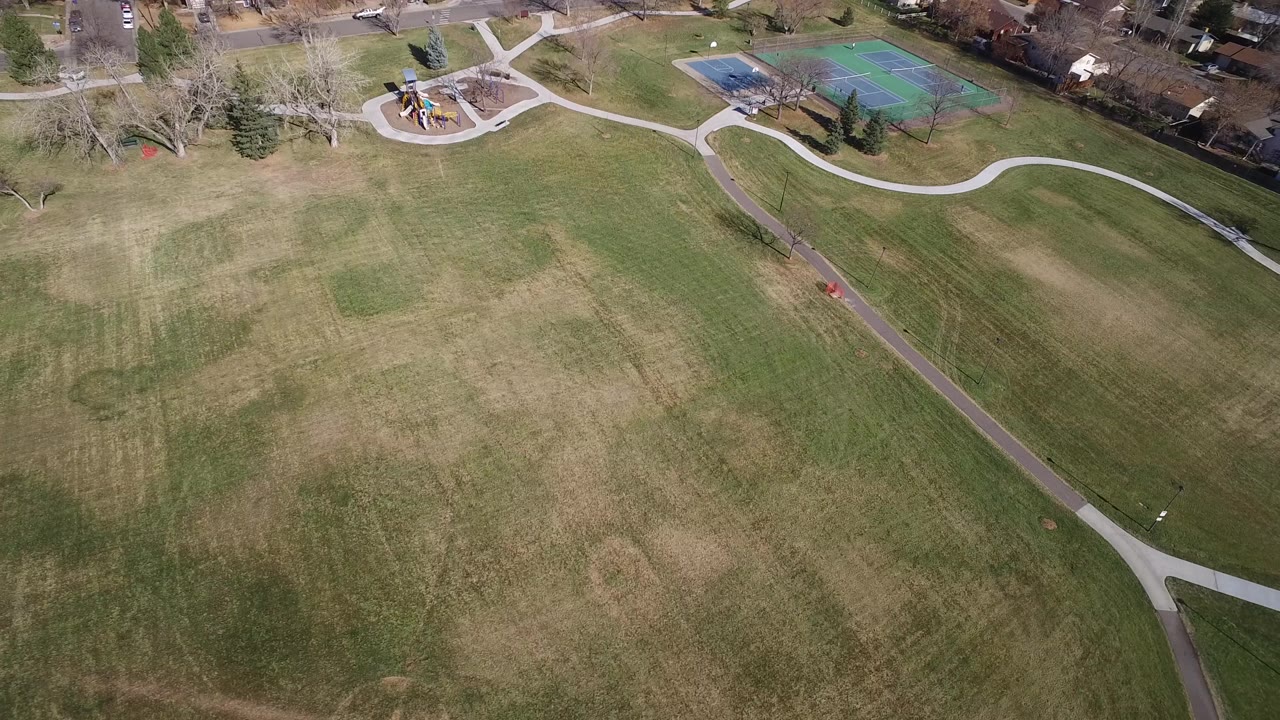 Aerial view in Westminster, Colorado showing patchy open grass below, a diagonal paved path, playground equipment and tennis courts above, with no visible people.