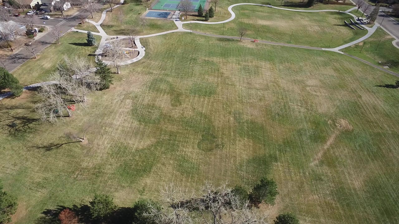 Aerial view of a large green field with curving paths and nearby tennis courts.