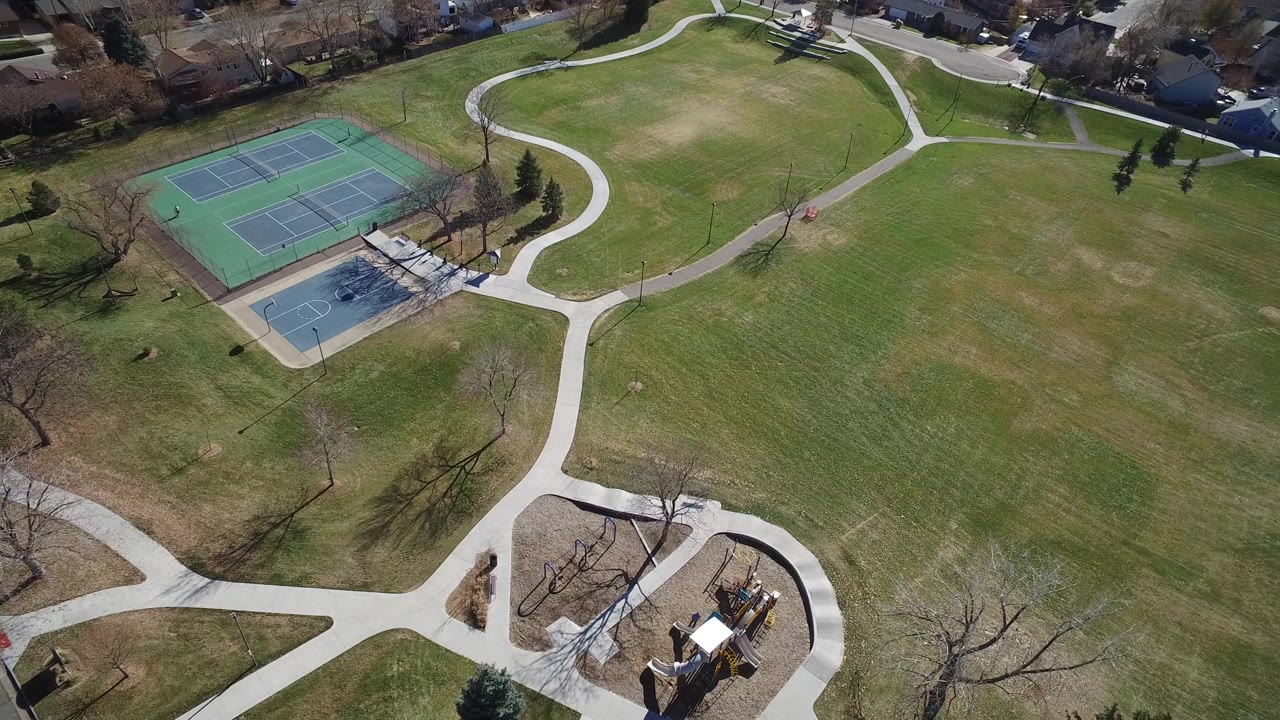 Overhead view of a Westminster, Colorado park with tennis courts at upper left, pale curving sidewalks, open grass, bare trees, playground equipment, and no visible people.