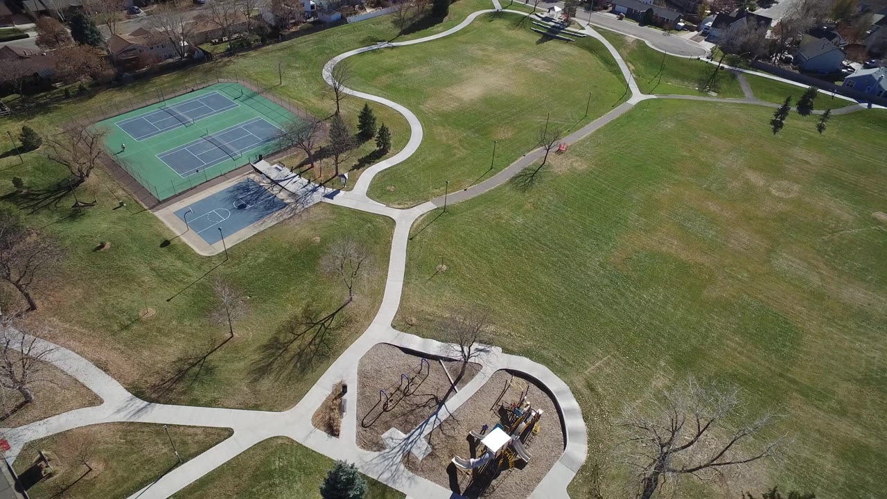 Aerial Westminster, Colorado park frame with tennis and basketball courts to the left, a large grass field across the center, a playground near the lower right, and no visible people.