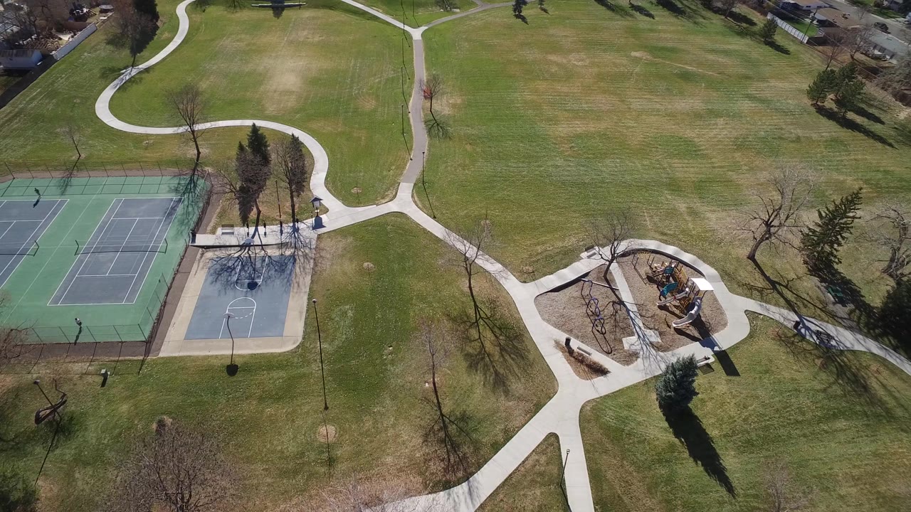 Overhead frame in Westminster, Colorado, with the fenced tennis courts more centered, a basketball half court below them, winding sidewalks, lawns, trees, and no visible people.