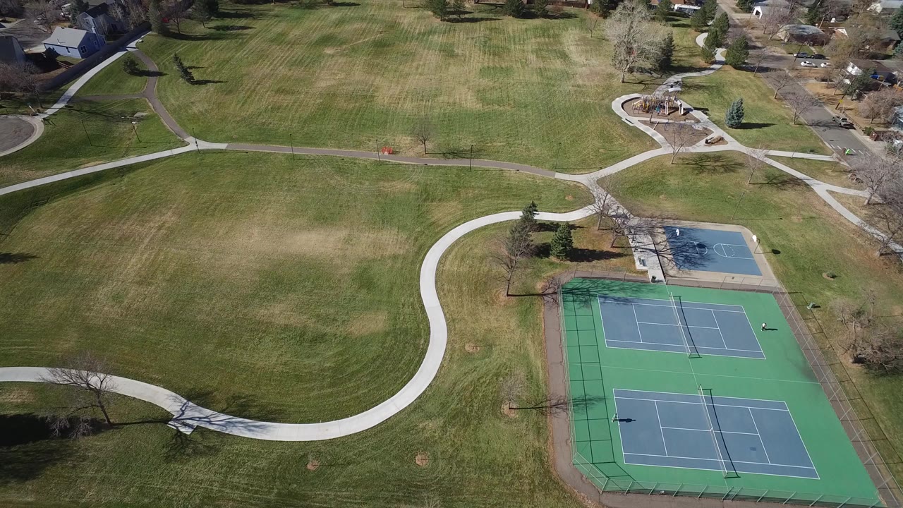 Aerial view over a Westminster, Colorado park with two green tennis courts, curving paths, open grass, leafless trees, nearby homes, and tiny distant figures possible near the courts.