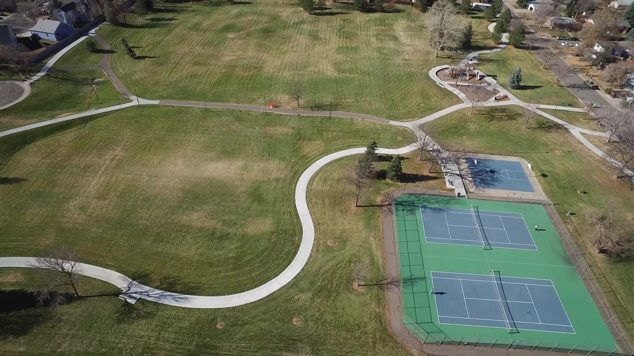 Drone frame over Westminster, Colorado showing tennis courts near the lower right, pale walking paths through mowed grass, bare trees, and residential streets beyond the park.