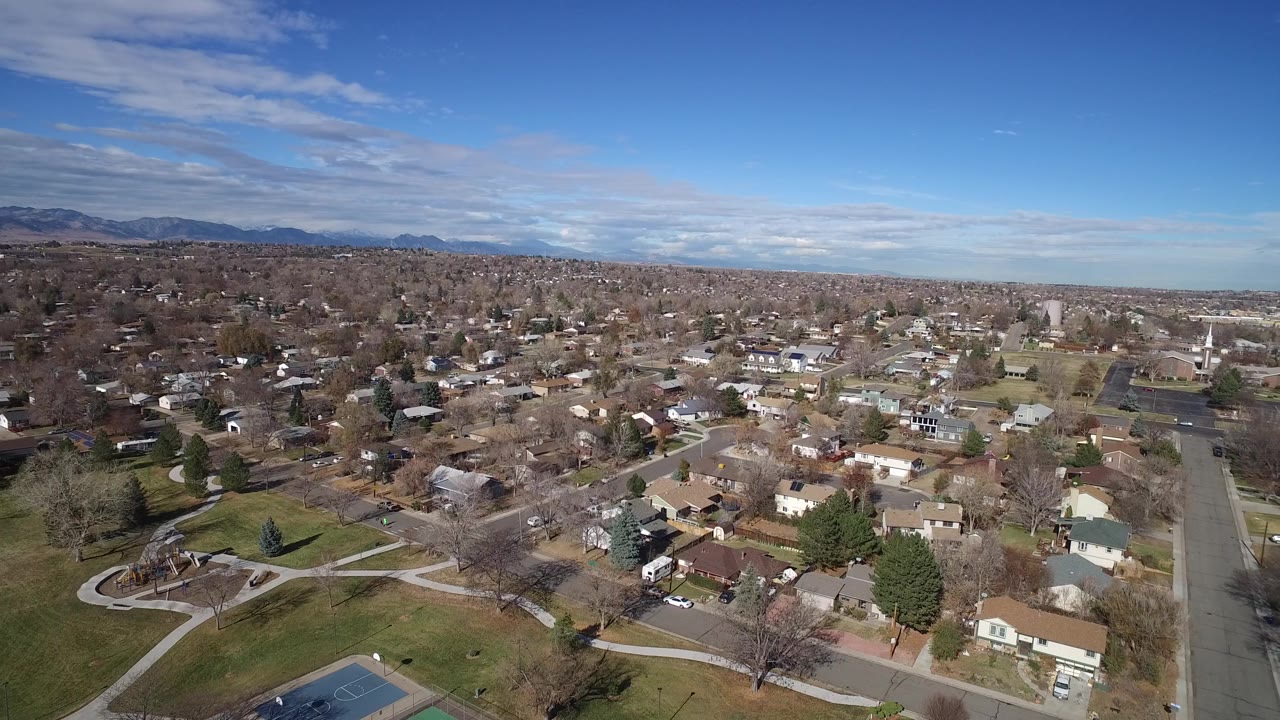 Aerial frame over Westminster, Colorado showing detached homes, a park edge with curved paths, a quiet road, and distant mountains beneath scattered clouds.