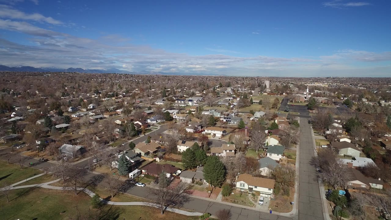Opening aerial frame over Westminster, Colorado, showing rooftops, a straight residential street, bare trees, distant mountains, blue sky, and no visible people.