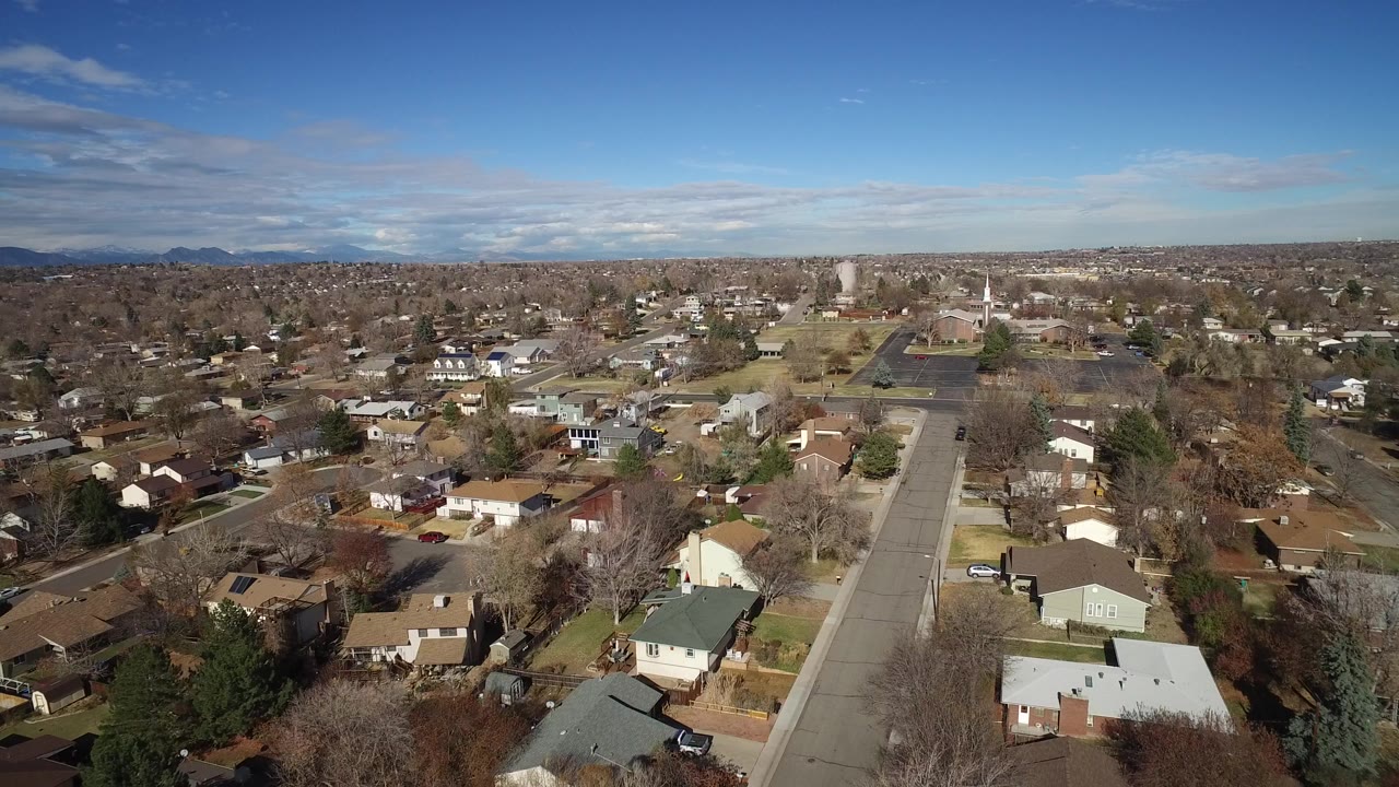 Wide aerial view of a Westminster, Colorado subdivision with houses, bare trees, a straight street running into the frame, distant mountains, and no visible people.