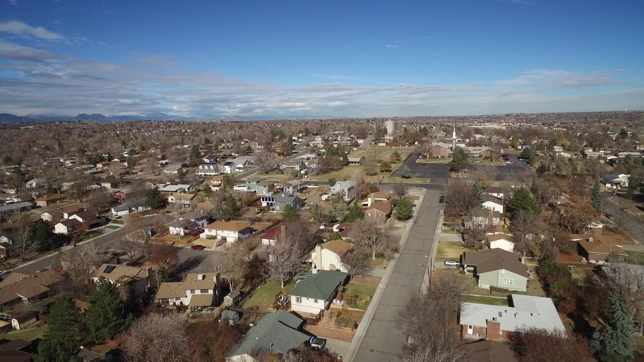Aerial frame over Westminster, Colorado homes with a large paved lot and white steeple-like structure in the midground, distant mountains, and no visible people.