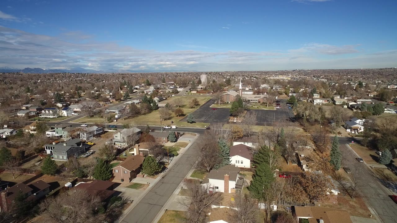 Aerial frame over Westminster, Colorado homes, residential roads, leafless trees, evergreen trees, distant mountains, and a broad blue sky, with no visible people.