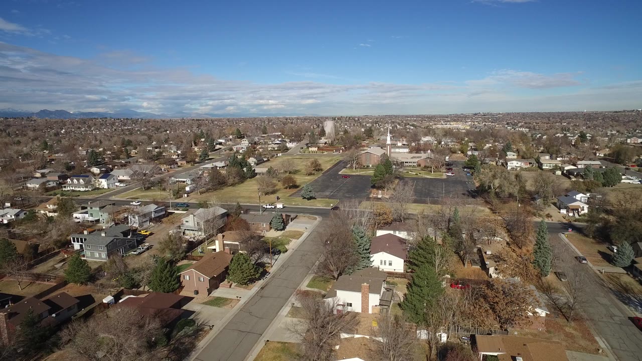 Overhead Westminster, Colorado view with houses in the foreground, a residential road, a large dark parking lot, scattered cars, trees, and no visible people.