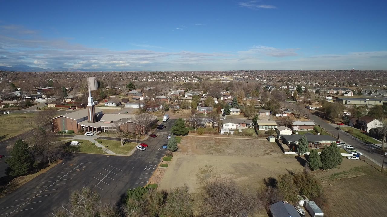 High aerial view over Westminster, Colorado, with tan open ground in front, a steepled brick building at left, houses and streets beyond, and no visible people.