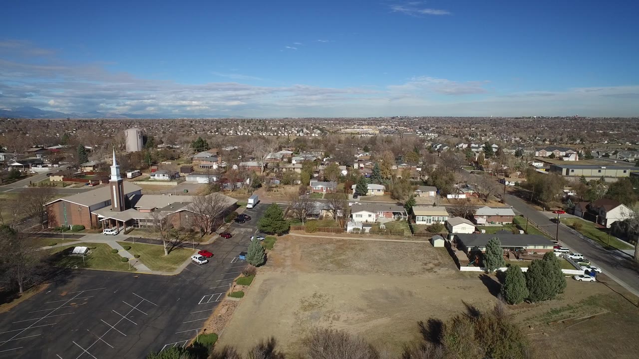 Aerial neighborhood view with a church-like steeple, open land, roads, and houses.